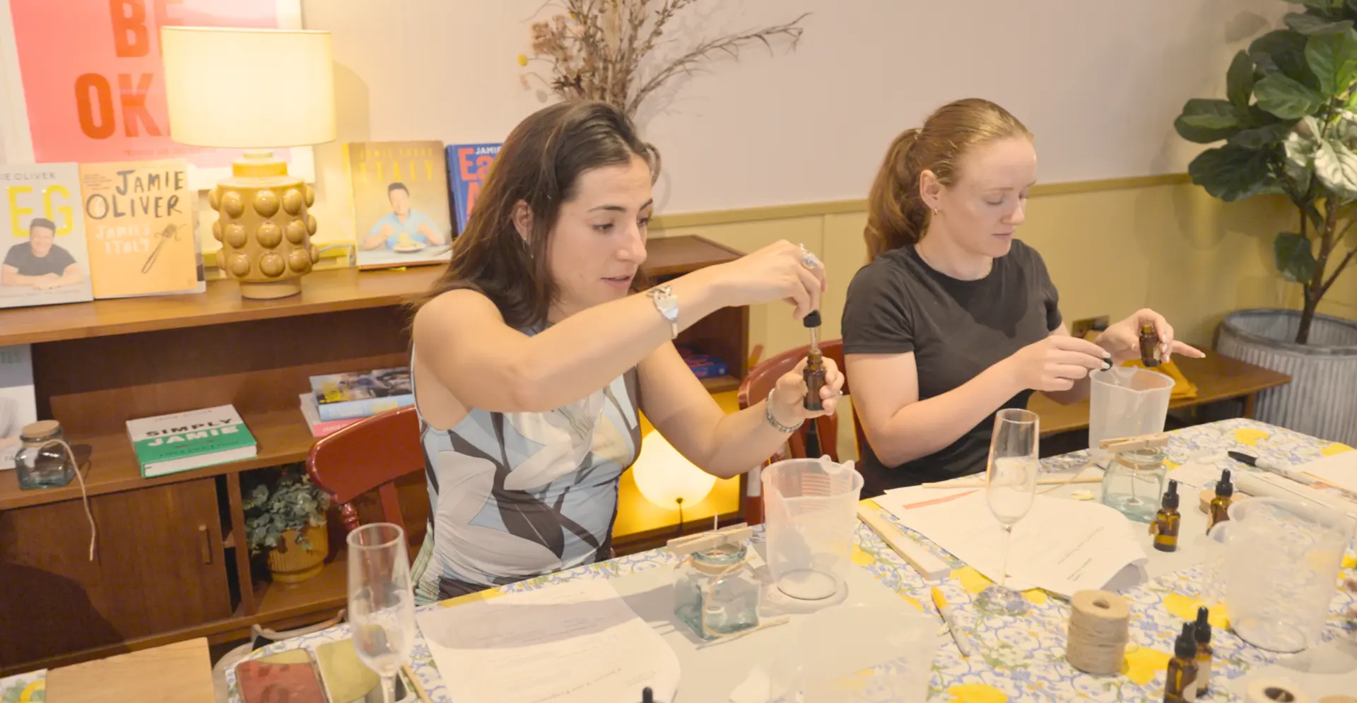 Two guests measuring fragrance oils with dropper bottles beside prepared candle jars